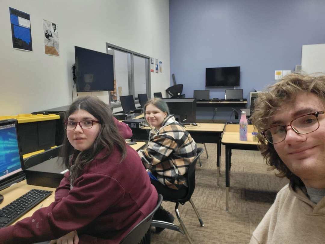 Rio taking the picture along with a girl with black hair in a red hoodie with glasses, and another girl with cyan hair, wearing plaid, are sitting in front of computers with Rio. Rio is holding the phone that took the photo of the group. This was during the Silver rank CyberPatriots semi-finals in 2023.