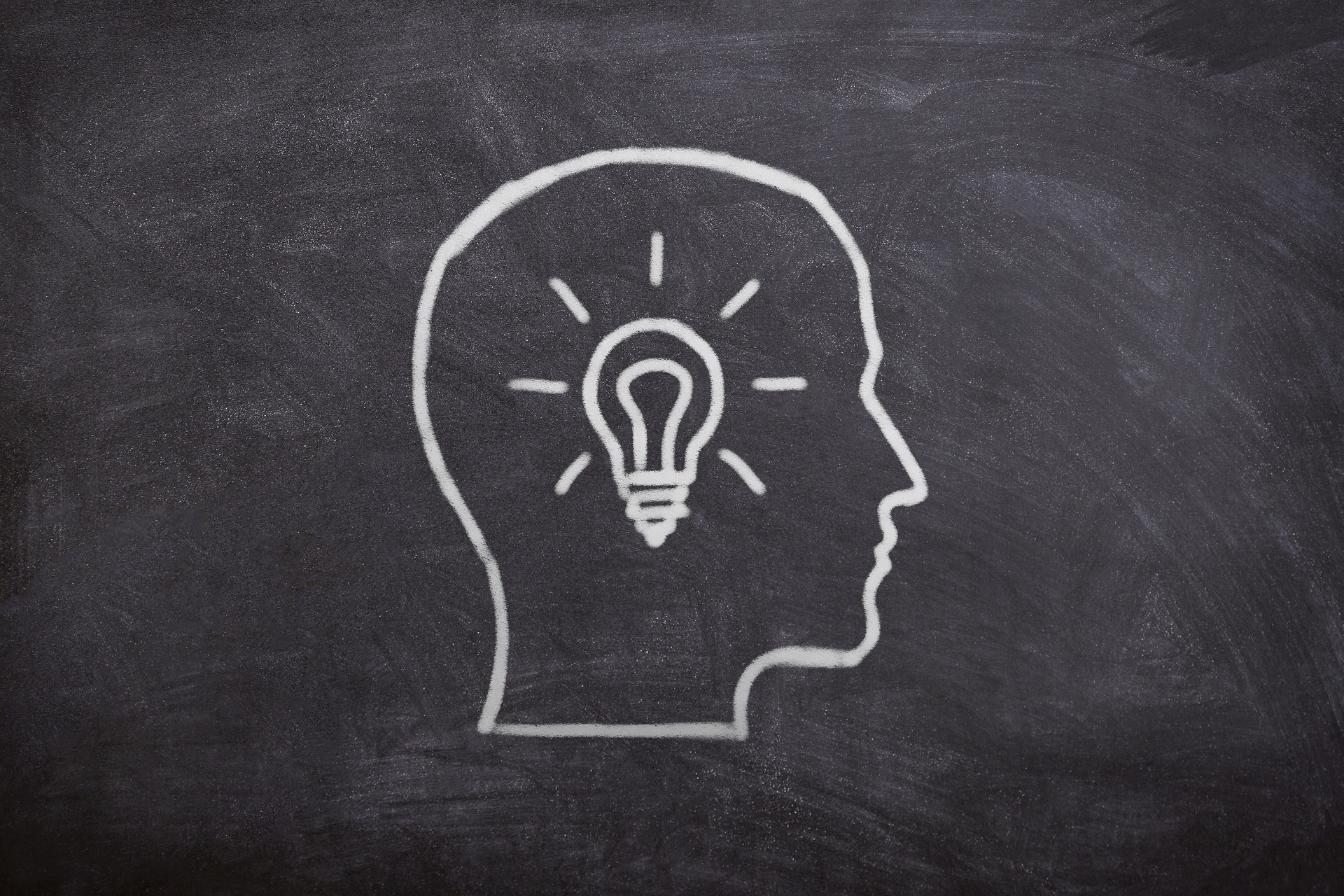 Drawing of a head of a person facing sideways in white on a chalkboard with a drawing of a lit up light bulb in yellow inside.