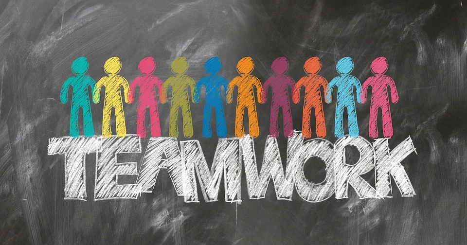 Multiple different colored stick figures drawn on a school chalkboard, holding hands. The word teamwork, in block letters, is drawn in white chalk underneath.